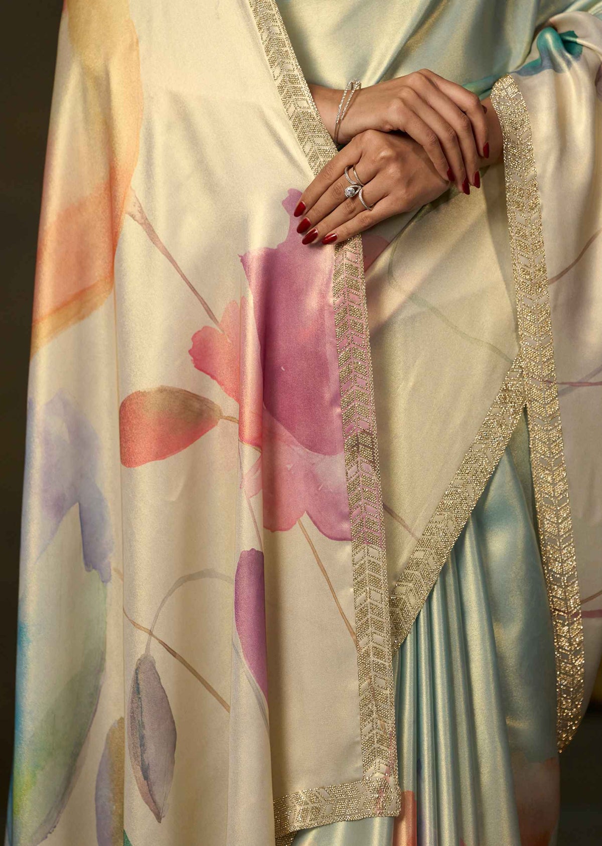A close-up of a draped satin fabric with a watercolor floral print in pink, purple, and peach, featuring a shimmering gold embroidered border. A person's hands with red nail polish and a silver ring rest on the fabric.