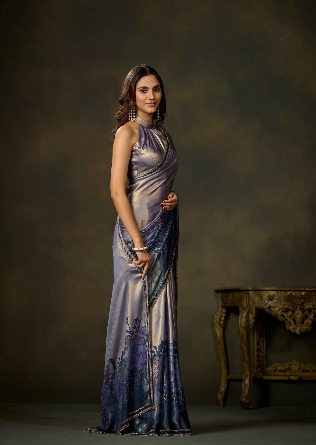 A woman modeling a sleeveless, floor-length metallic sari in a studio setting. The sari has a shimmering lavender-purple body with a darker blue and purple floral pattern near the lower hem.