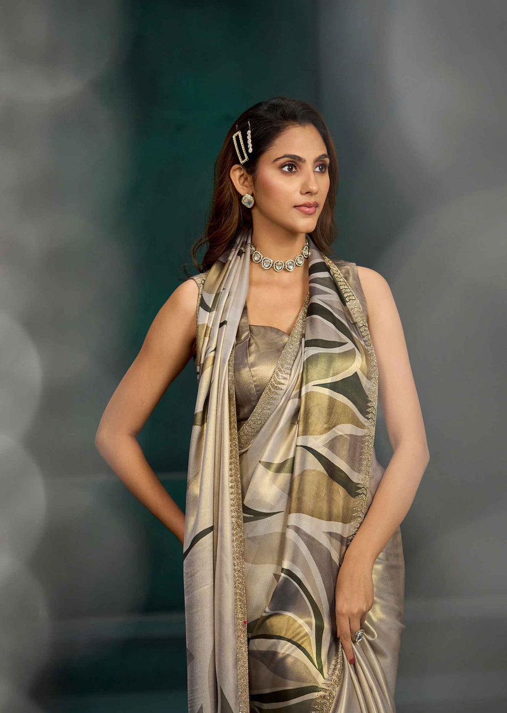 A woman models a sleeveless metallic gold blouse paired with a beige and olive green patterned saree and dupatta, set against a dark gray bokeh background.