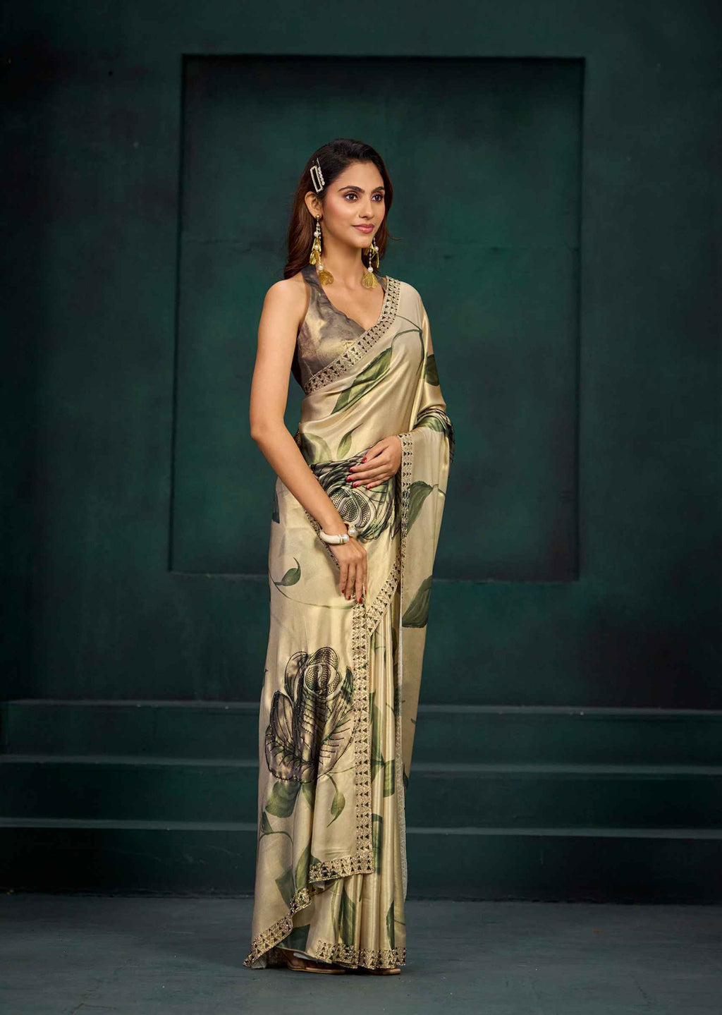 A woman modeling a beige saree with green and black floral prints, paired with a matching sleeveless blouse, standing against a dark green studio backdrop.
