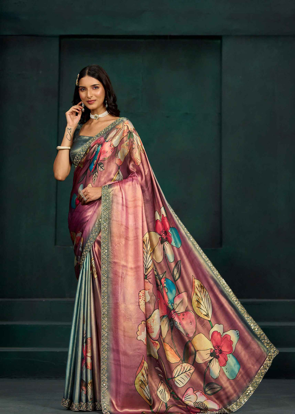 A woman models a traditional Indian saree with a dusty pink and purple body, decorated with a vibrant multicolor floral print on the pallu and border, set against a dark green textured studio backdrop.