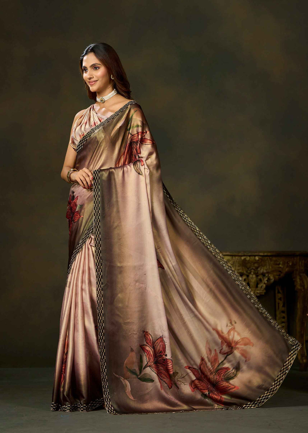 A woman modeling a shimmering champagne-gold saree with red floral motifs and a black patterned border, posed against a dark brown studio backdrop.