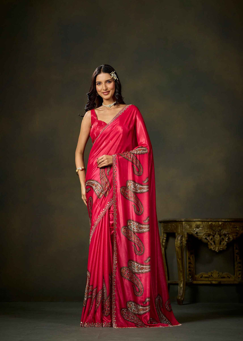 A woman modeling a bright pink saree with gold paisley embroidery, standing in front of a dark brown studio backdrop with an ornate table partially visible on the right.