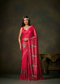 A woman modeling a bright pink saree with gold paisley embroidery, standing in front of a dark brown studio backdrop with an ornate table partially visible on the right.