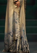 A close-up of a draped garment or fabric with a metallic champagne-beige base, watercolor-style blue and gray painterly print, and a black-and-beige patterned border, shown on a model against a dark green background.