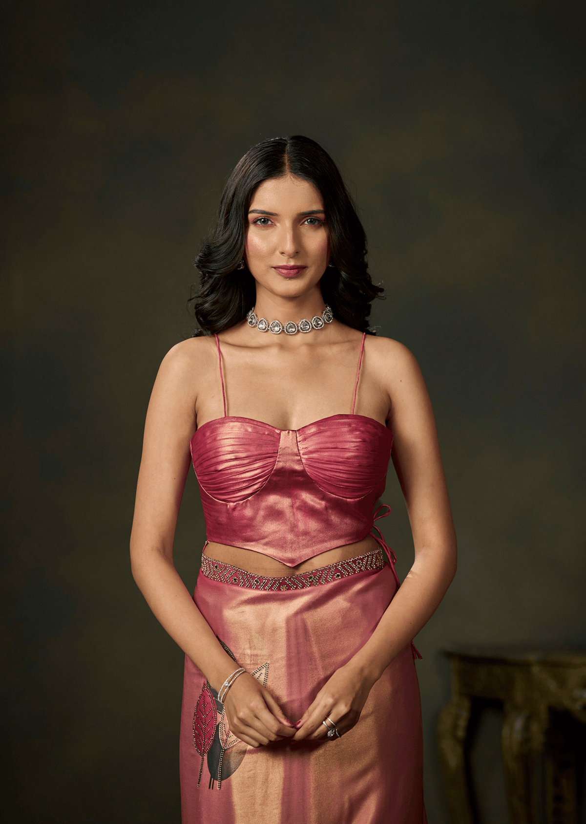 A woman wearing a shiny pink two-piece outfit with a ruched sweetheart-style top and a matching skirt, posed against a dark brown studio background.