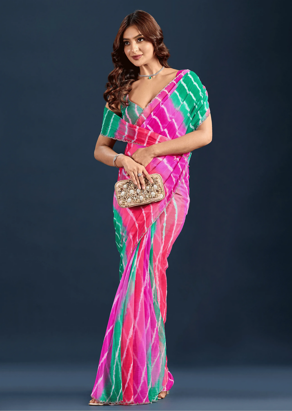 A woman modeling a vibrant pink and green tie-dye saree with a matching blouse, holding a small metallic clutch against a dark blue studio background.