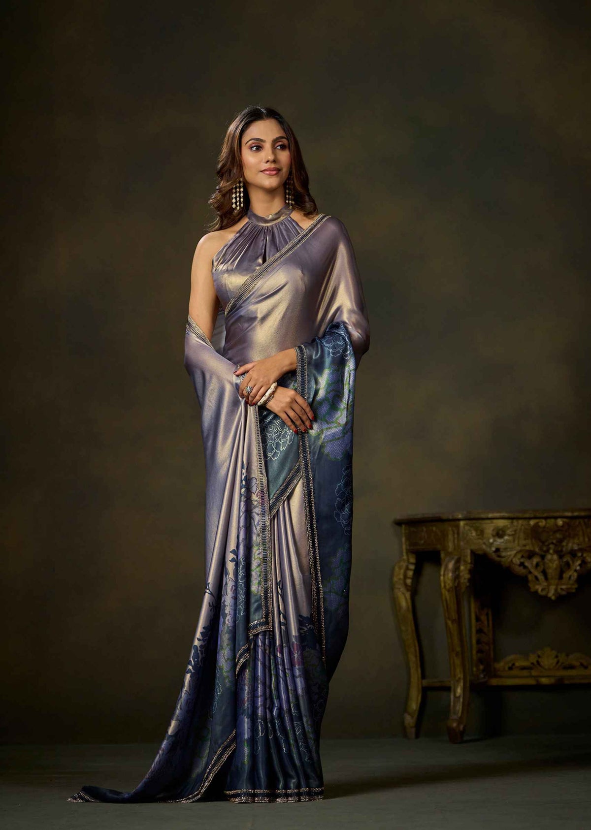 A woman models a floor-length draped gown with a metallic lavender-purple bodice and a blue patterned skirt, standing against a dark brown studio backdrop with an ornate table to the side.