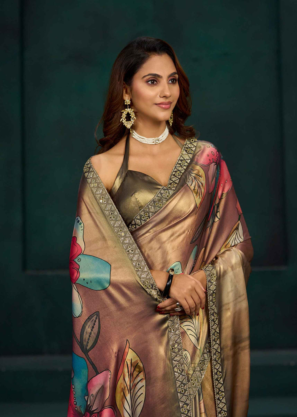A woman wearing a metallic bronze and gold saree with colorful floral prints in pink, teal, and green, paired with a matching metallic blouse, set against a dark green background.