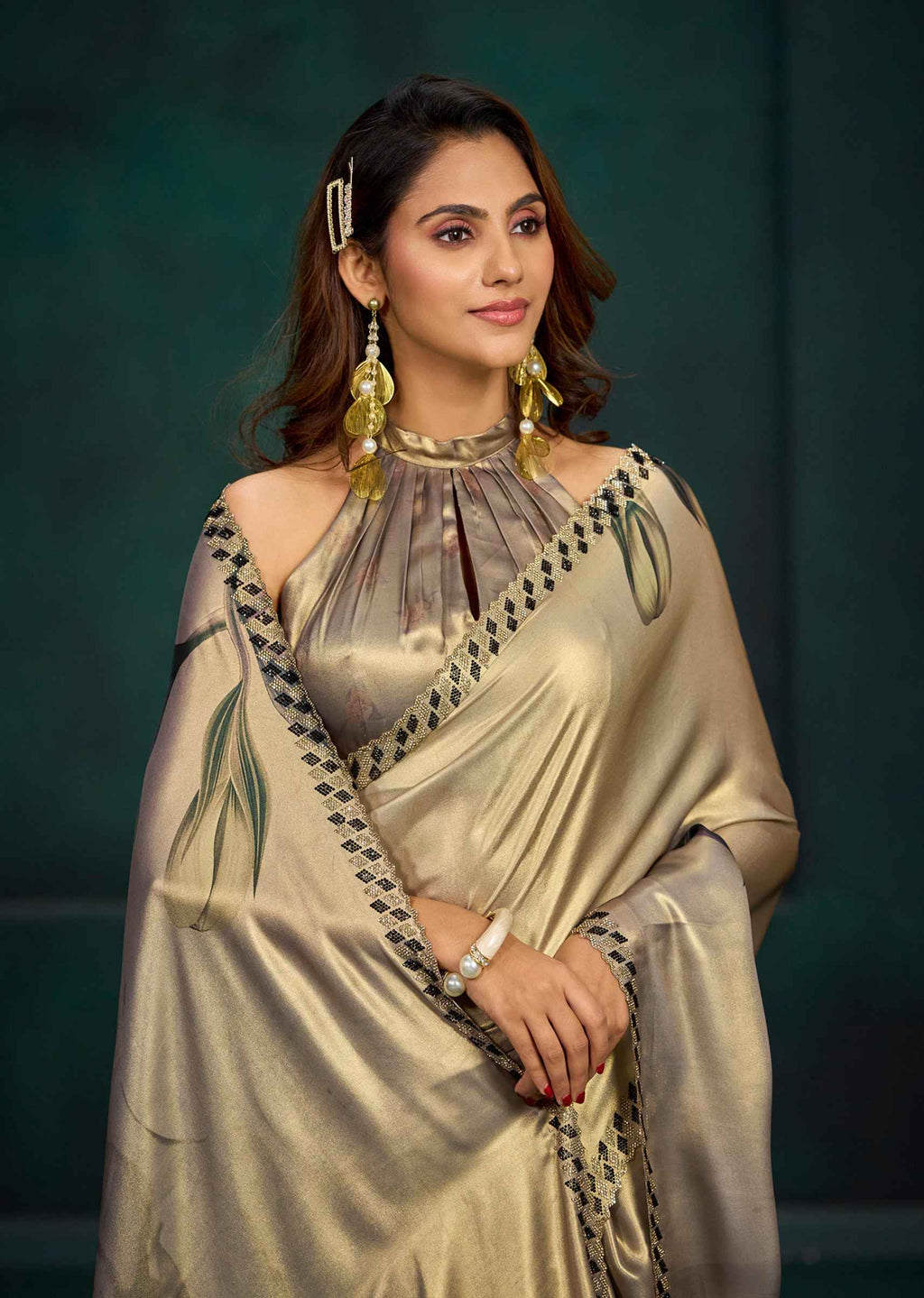 A woman modeling a champagne gold satin saree with black and green border detailing, paired with a matching metallic blouse, against a dark green studio backdrop.