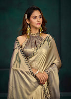 A woman modeling a champagne gold satin saree with black and green border detailing, paired with a matching metallic blouse, against a dark green studio backdrop.