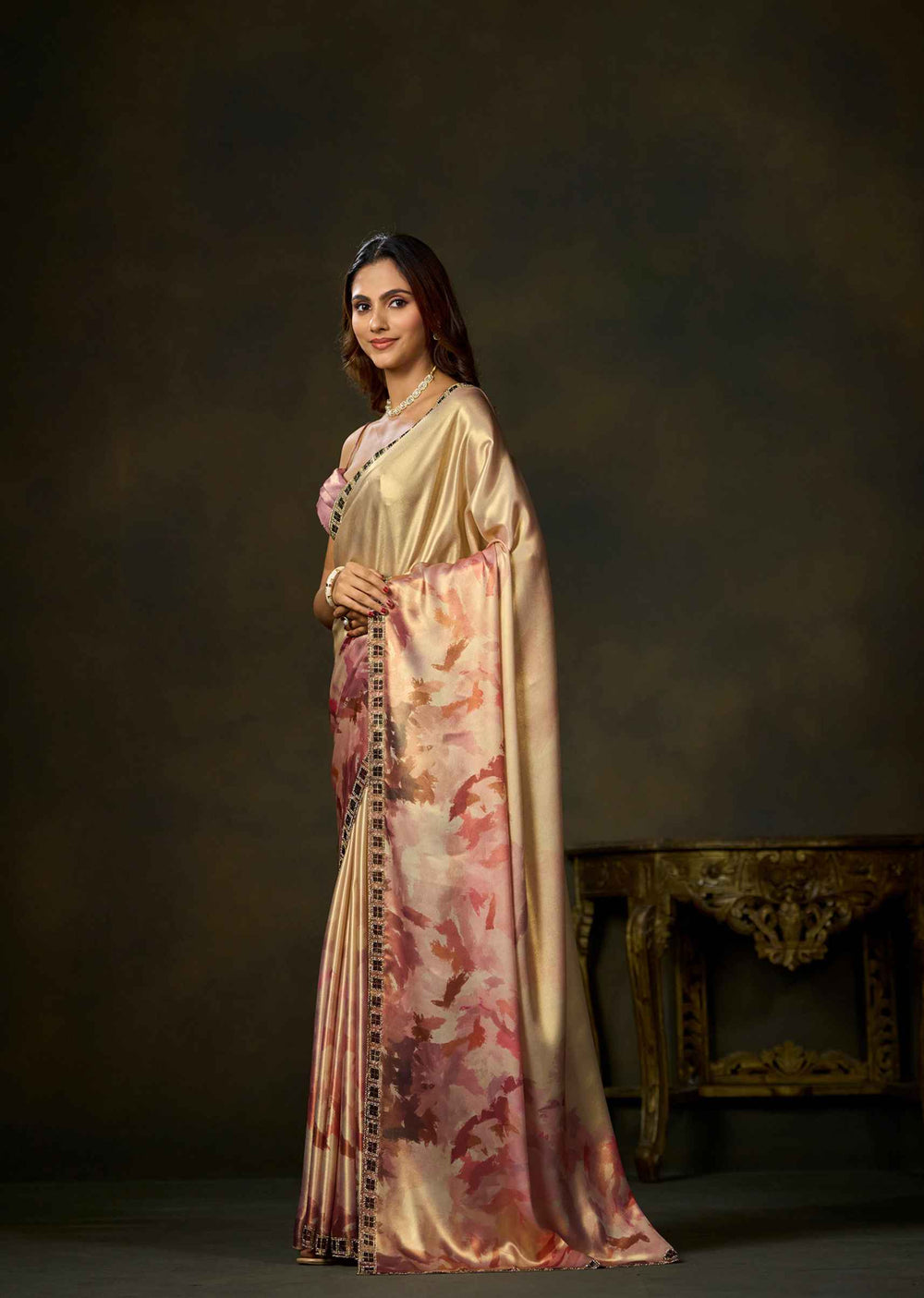 A woman modeling a traditional Indian saree in a studio setting. The saree has a shiny beige-gold body with a pink and red floral print on the lower portion and a dark patterned border.