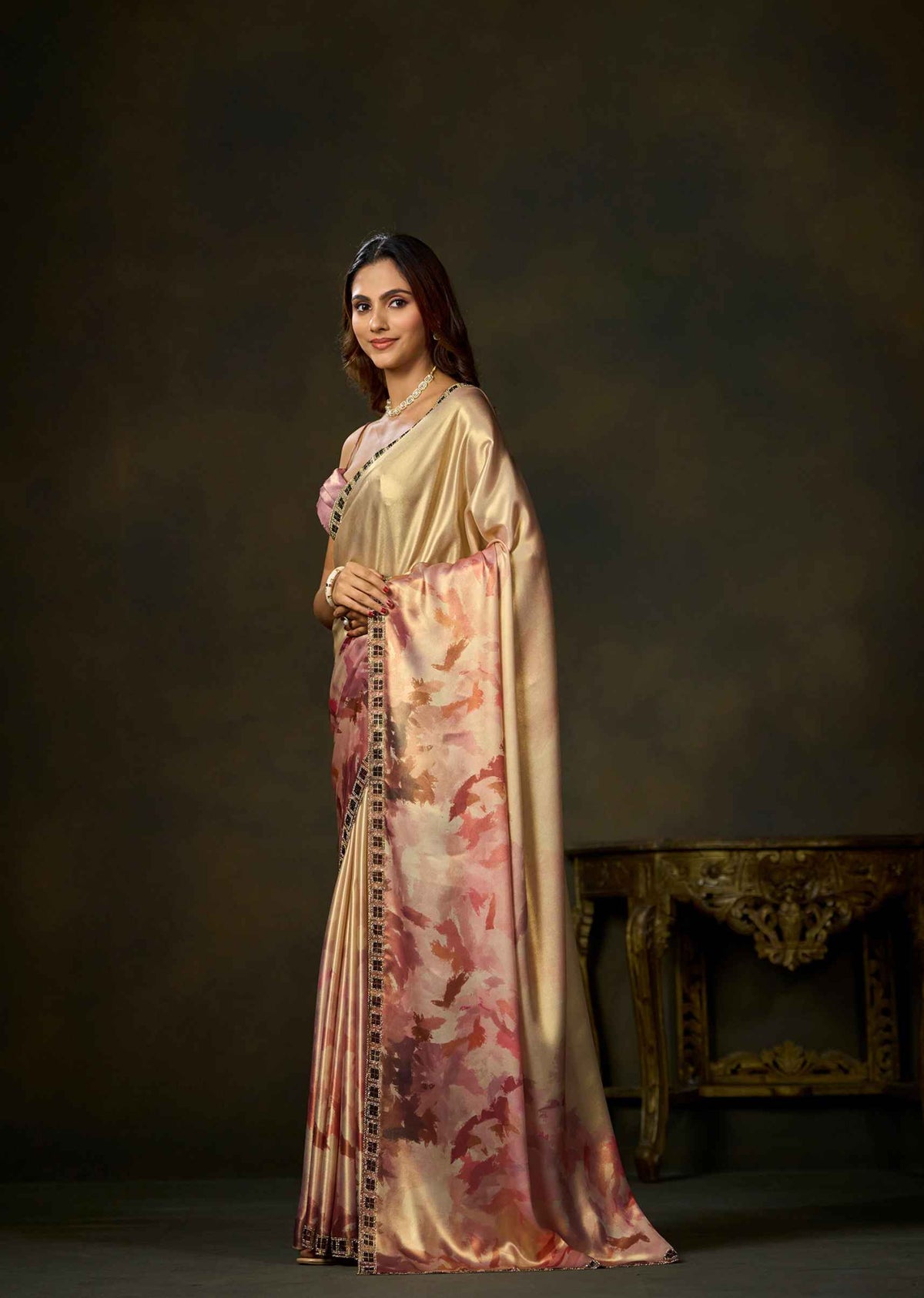A woman modeling a traditional Indian saree in a studio setting. The saree has a shiny beige-gold body with a pink and red floral print on the lower portion and a dark patterned border.