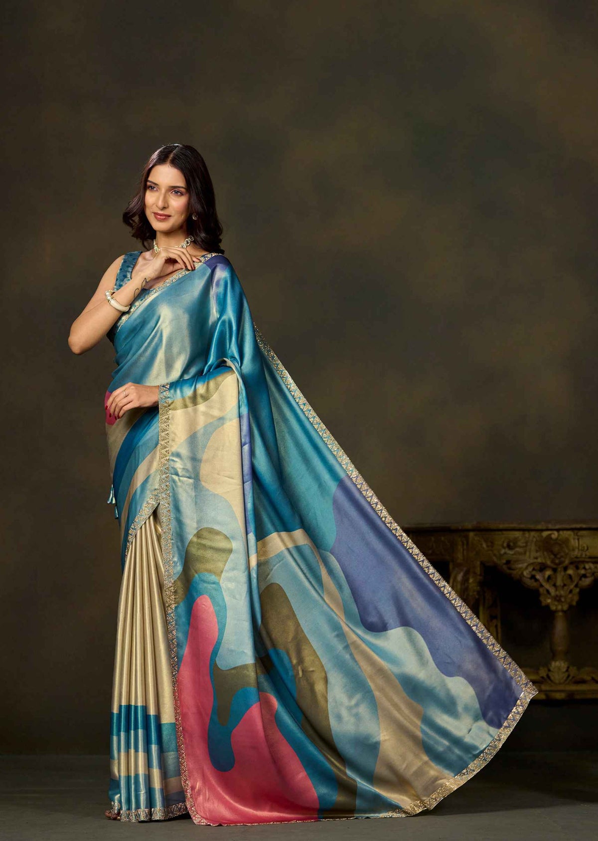 A woman models a luxurious blue and beige silk saree with abstract pink, blue, and gold patterns, posed against a dark brown studio backdrop.