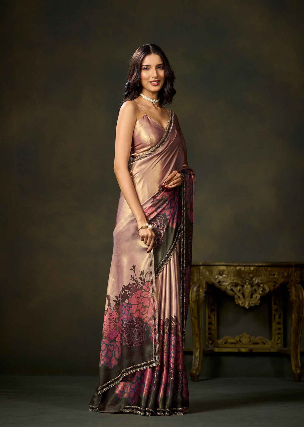 A woman modeling a draped satin saree with a metallic rose gold base and dark pink floral patterns near the hem, standing in front of a dark brown studio backdrop with an ornate gold table.