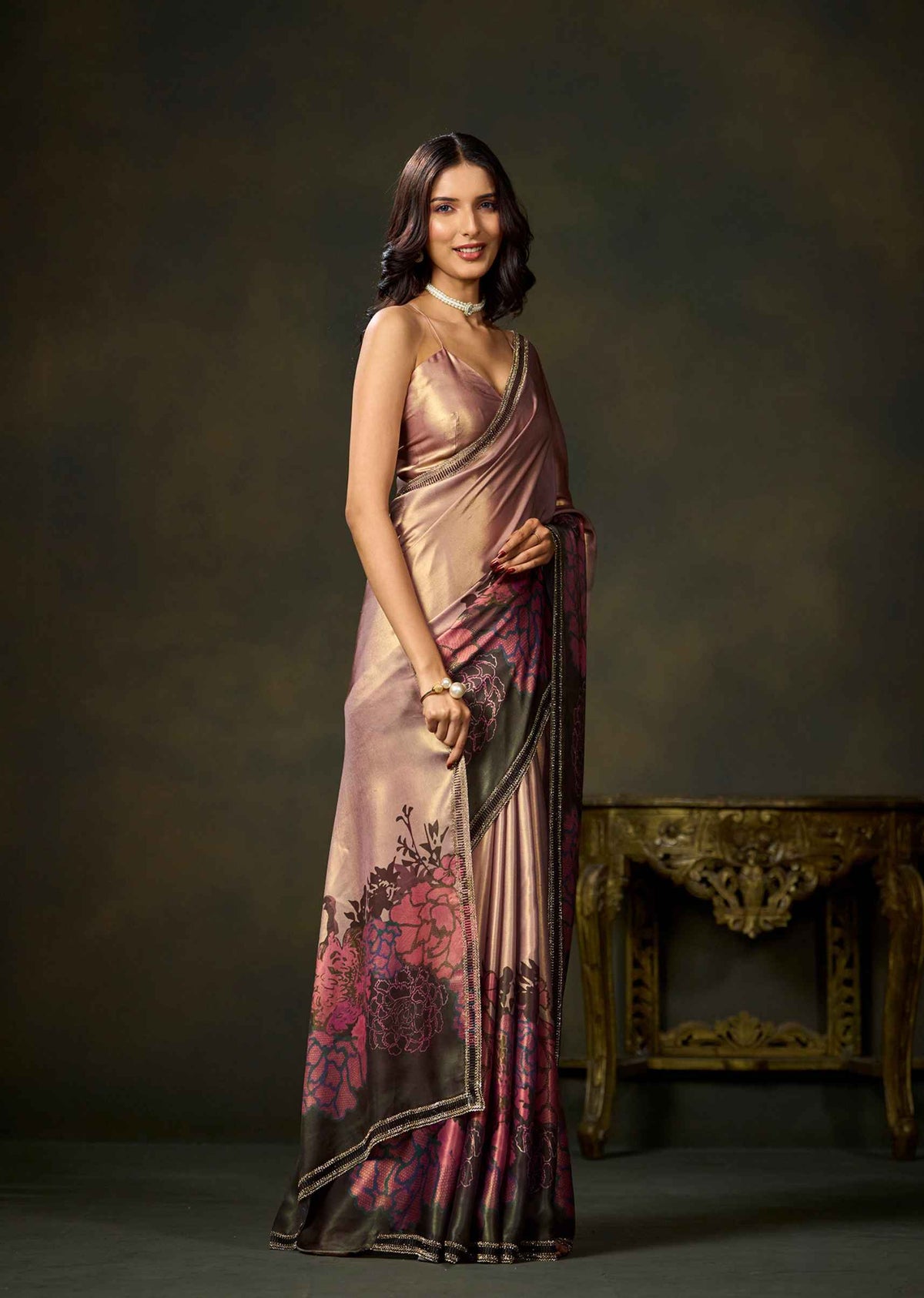 A woman modeling a draped satin saree with a metallic rose gold base and dark pink floral patterns near the hem, standing in front of a dark brown studio backdrop with an ornate gold table.