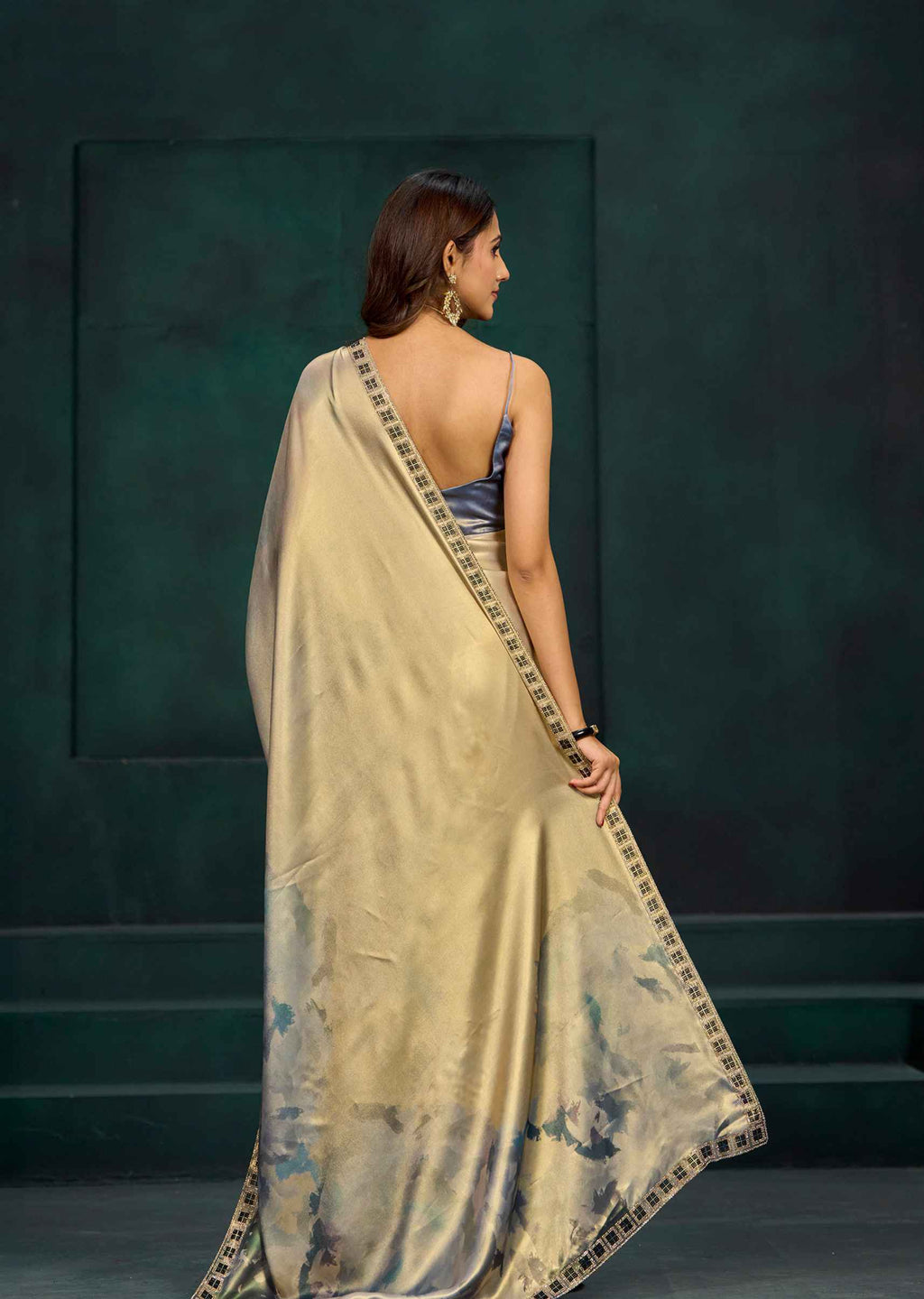 A woman is shown from behind wearing a beige silk saree with a blue and black patterned border, paired with a blue sleeveless blouse, set against a dark green textured studio background.