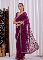 A woman models a deep purple georgette saree with intricate silver floral embroidery and a matching embroidered dupatta, set against a white curtain backdrop with a decorative wooden screen on the left.