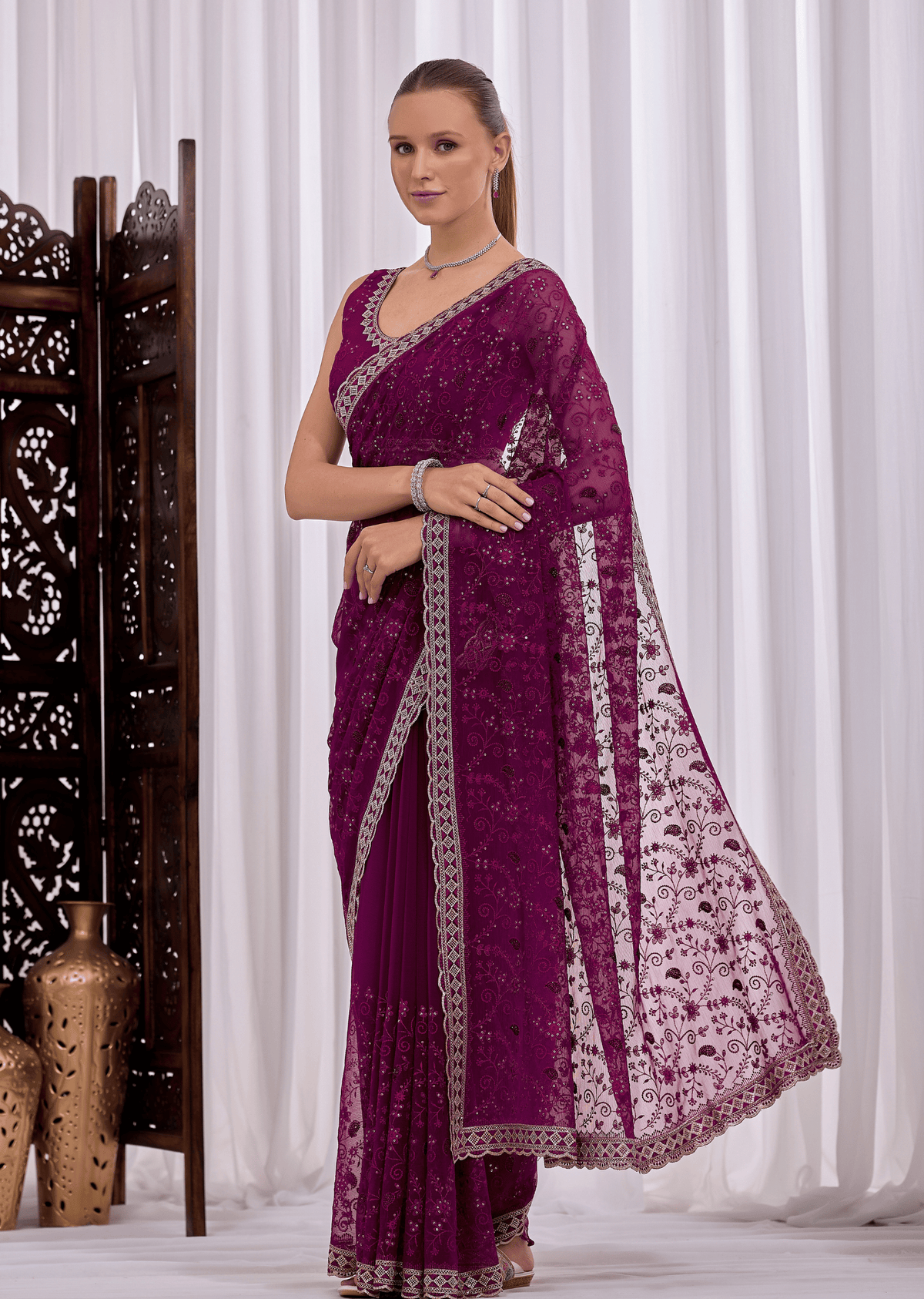 A woman models a deep purple georgette saree with intricate silver floral embroidery and a matching embroidered dupatta, set against a white curtain backdrop with a decorative wooden screen on the left.