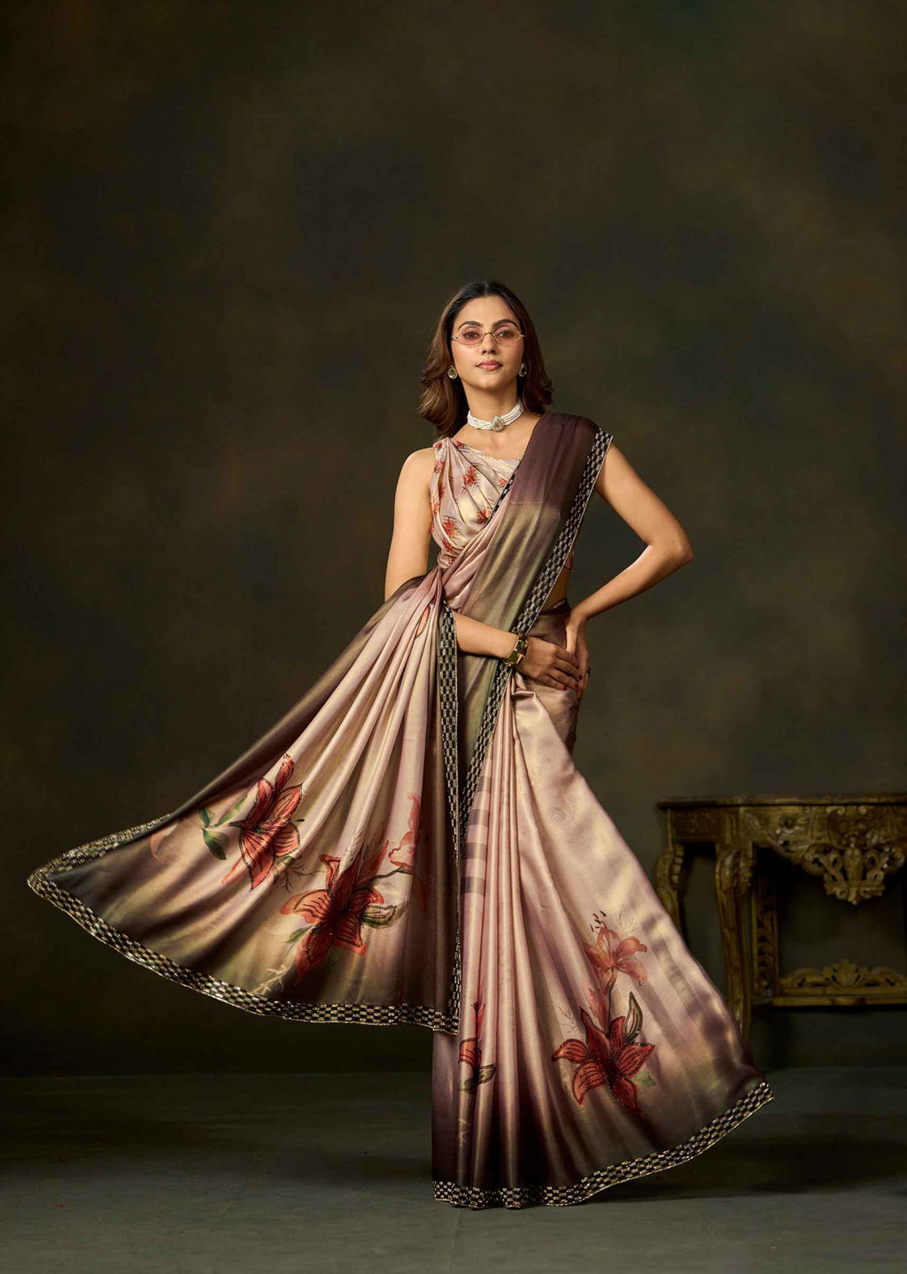 A woman models a draped satin saree with a metallic rose gold and bronze sheen, decorated with red floral motifs and a black and gold patterned border, set against a dark brown studio background.