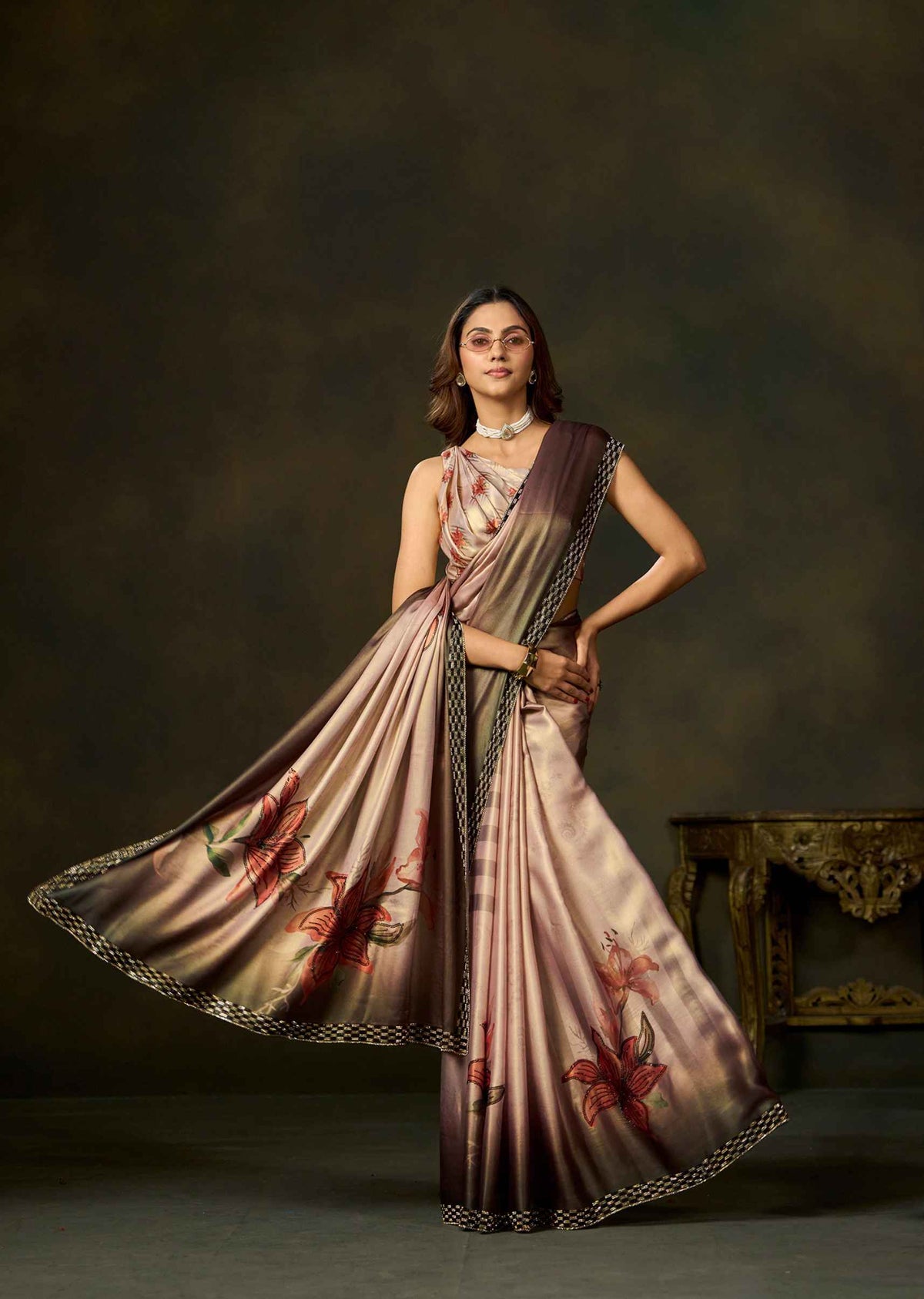 A woman models a draped satin saree with a metallic rose gold and bronze sheen, decorated with red floral motifs and a black and gold patterned border, set against a dark brown studio background.