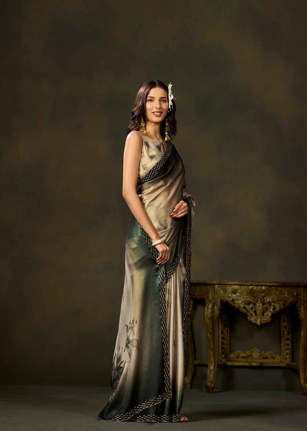 A woman models a sleeveless, floor-length draped dress with a metallic champagne-gold and dark green gradient, featuring black patterned borders. She is standing in a studio setting with a dark brown textured backdrop and an ornate gold table on the right.