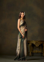 A woman models a sleeveless, floor-length draped dress with a metallic champagne-gold and dark green gradient, featuring black patterned borders. She is standing in a studio setting with a dark brown textured backdrop and an ornate gold table on the right.