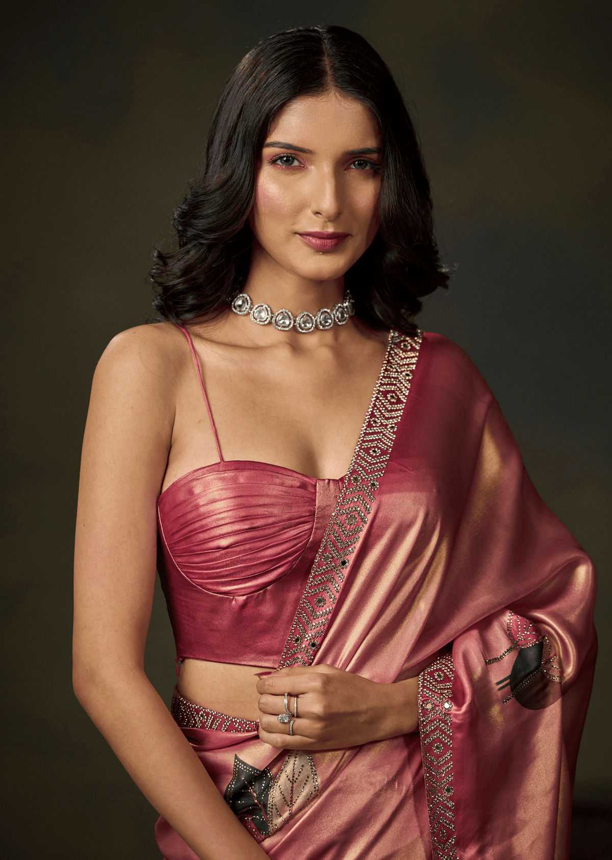 A woman modeling a metallic pink saree with a matching pink blouse and a silver choker necklace, set against a dark, neutral studio background.