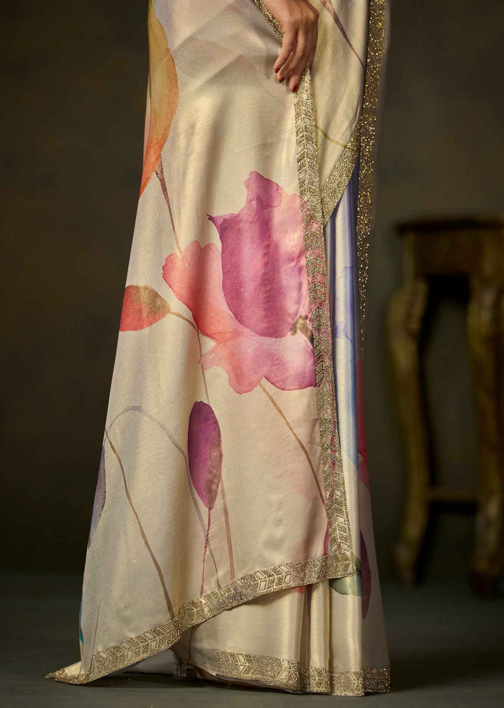 A close-up of a draped saree with a cream base, large pink and purple floral print, and a gold embroidered border, shown on a model in a dimly lit indoor setting.