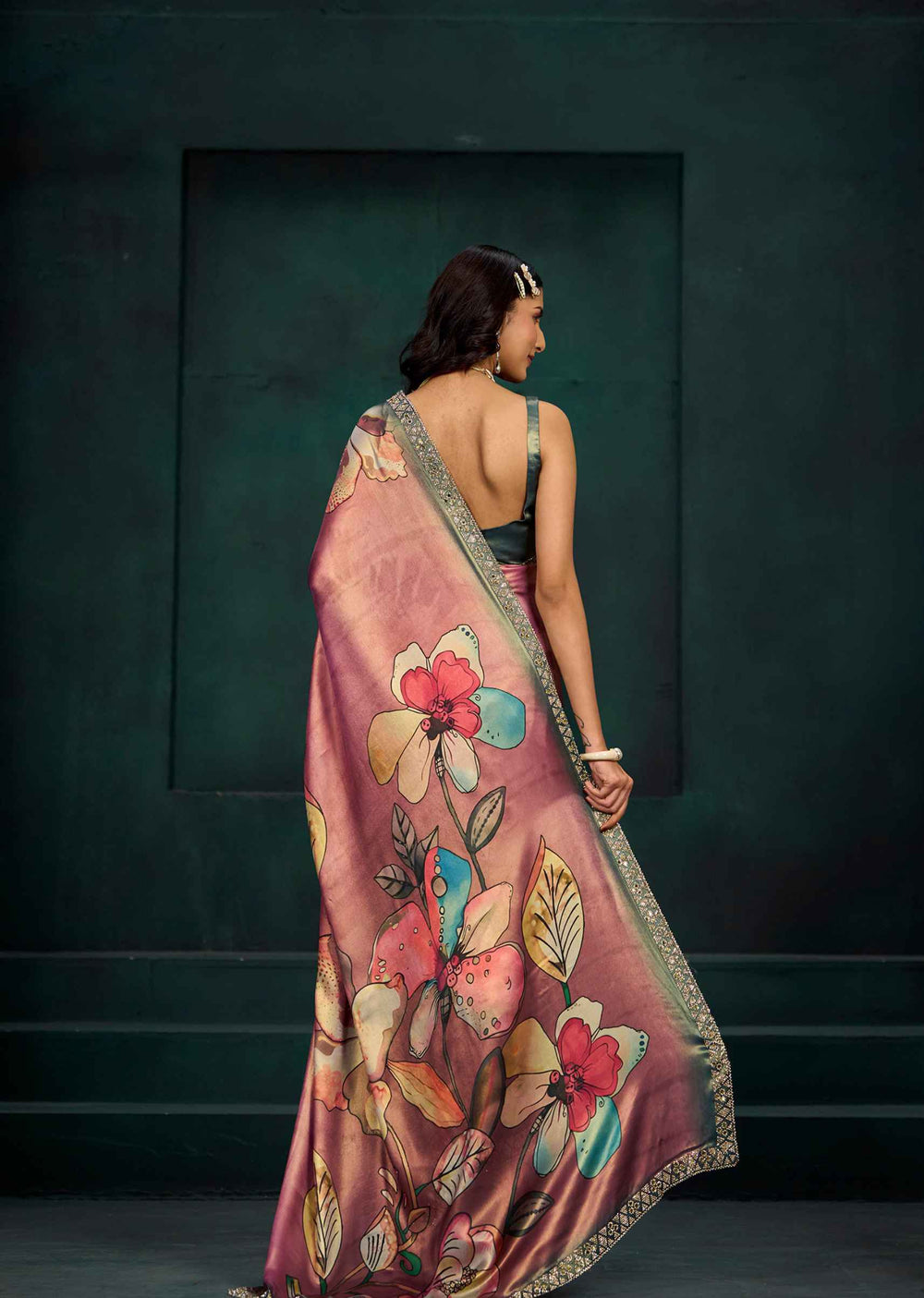 A woman is shown from behind wearing a pink floral saree with a metallic gold border, set against a dark teal textured wall.