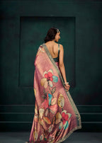 A woman is shown from behind wearing a pink floral saree with a metallic gold border, set against a dark teal textured wall.