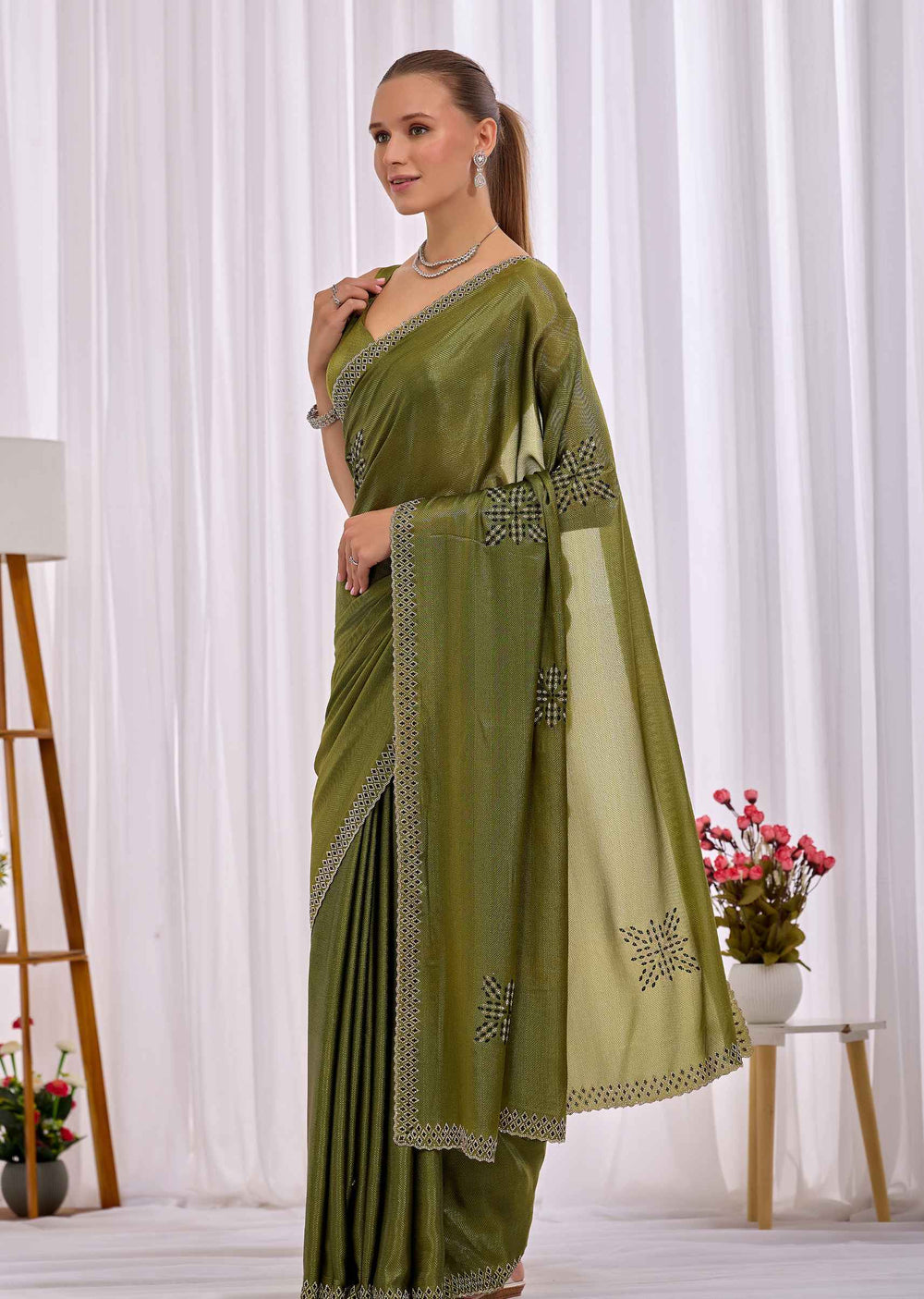 A woman modeling an olive green saree with silver embroidered borders and motifs, standing in front of a white curtain backdrop.