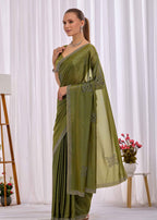 A woman modeling an olive green saree with silver embroidered borders and motifs, standing in front of a white curtain backdrop.