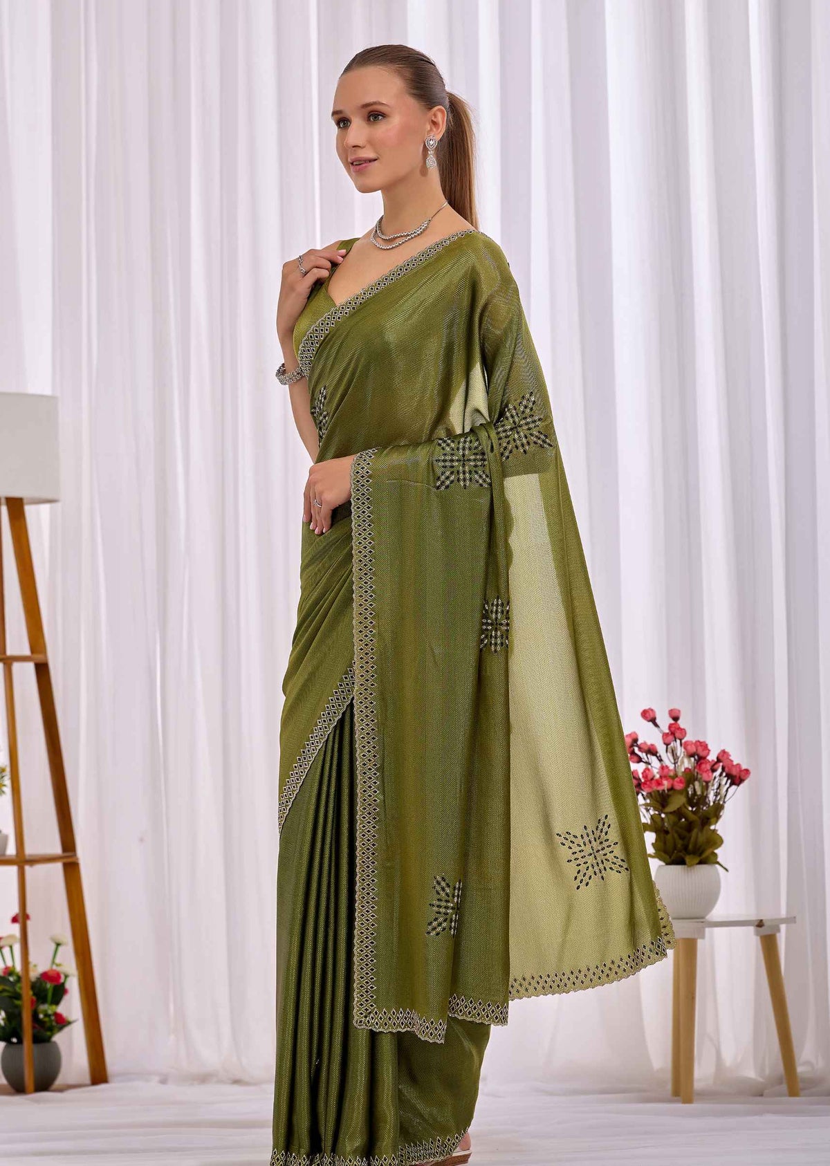 A woman modeling an olive green saree with silver embroidered borders and motifs, standing in front of a white curtain backdrop.