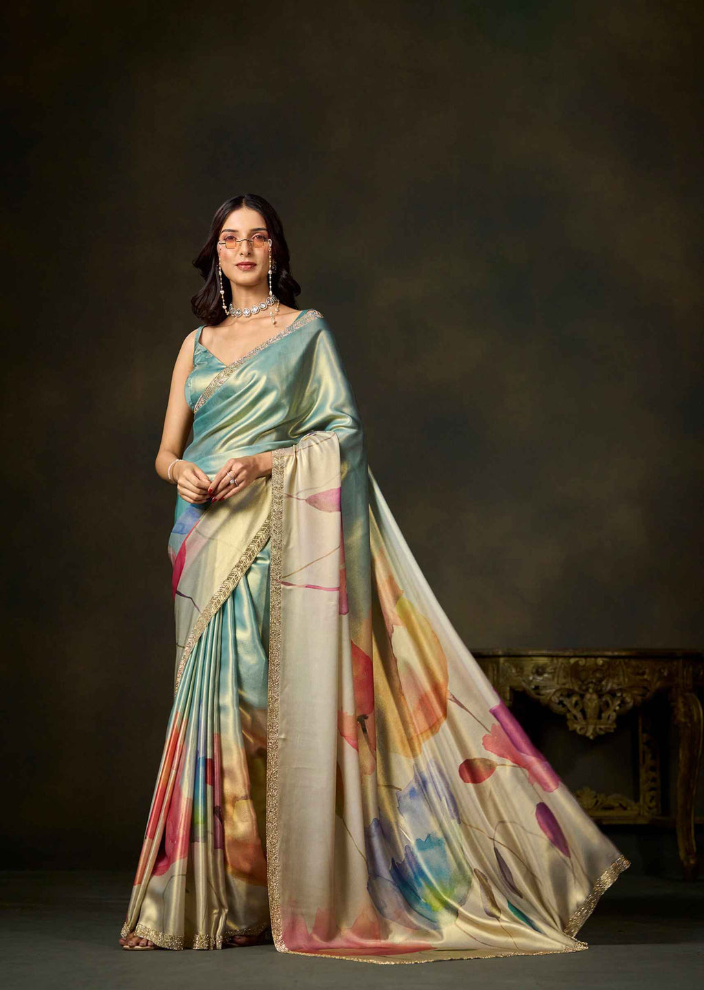A woman models a shimmering satin saree with a light blue-green body and a beige border decorated with colorful abstract watercolor-like prints in pink, orange, yellow, and blue. She is posed in front of a dark, moody studio background.