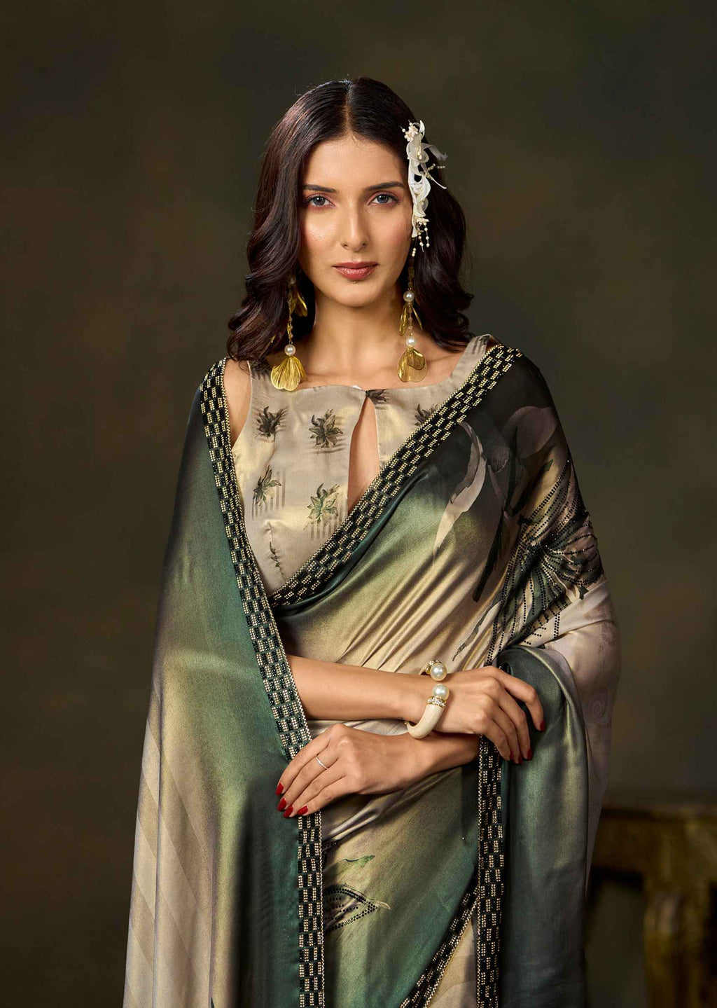 A woman modeling a traditional Indian saree with a green and beige floral print, paired with a matching dupatta and gold jewelry, set against a dark, textured studio background.