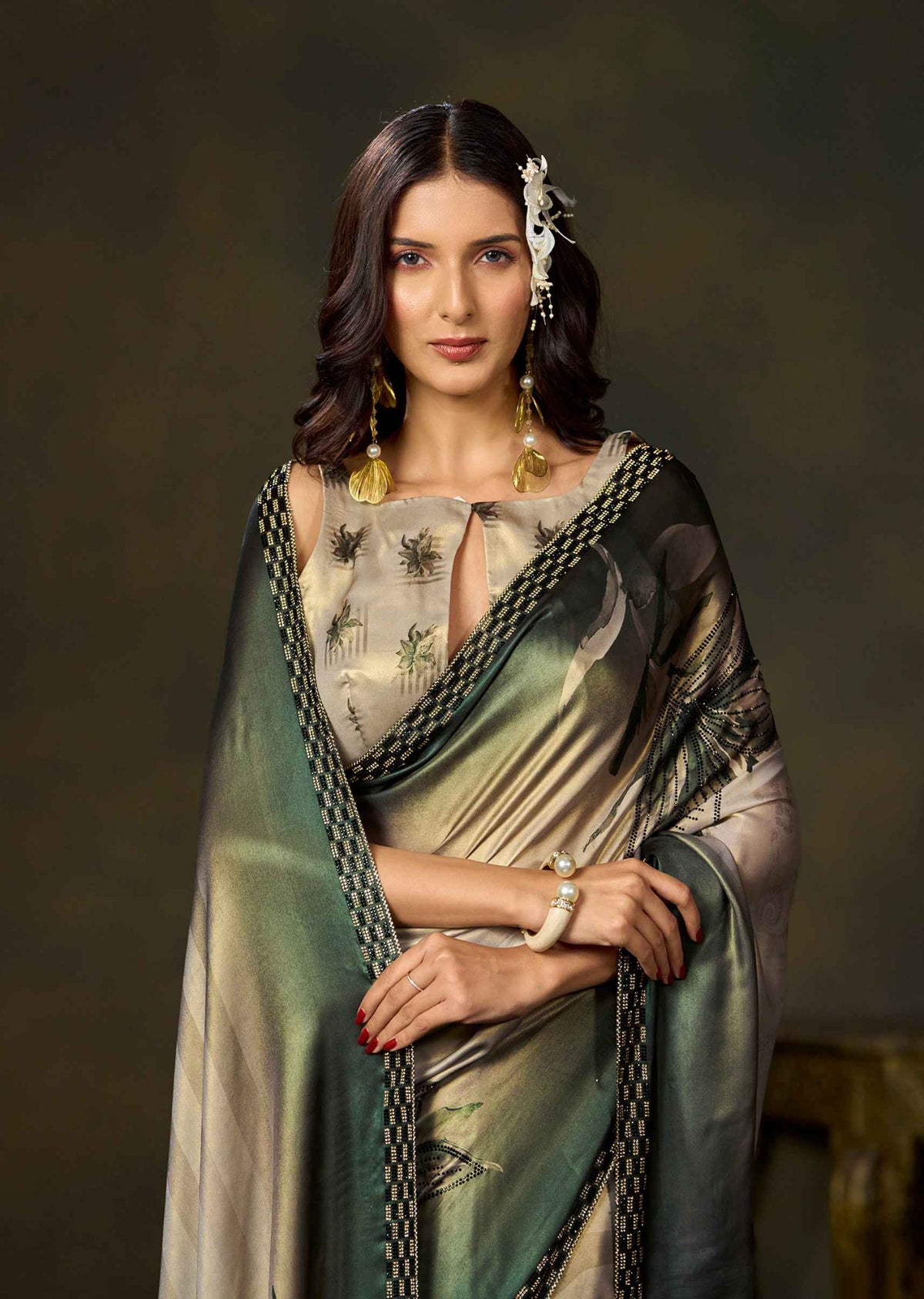 A woman modeling a traditional Indian saree with a green and beige floral print, paired with a matching dupatta and gold jewelry, set against a dark, textured studio background.