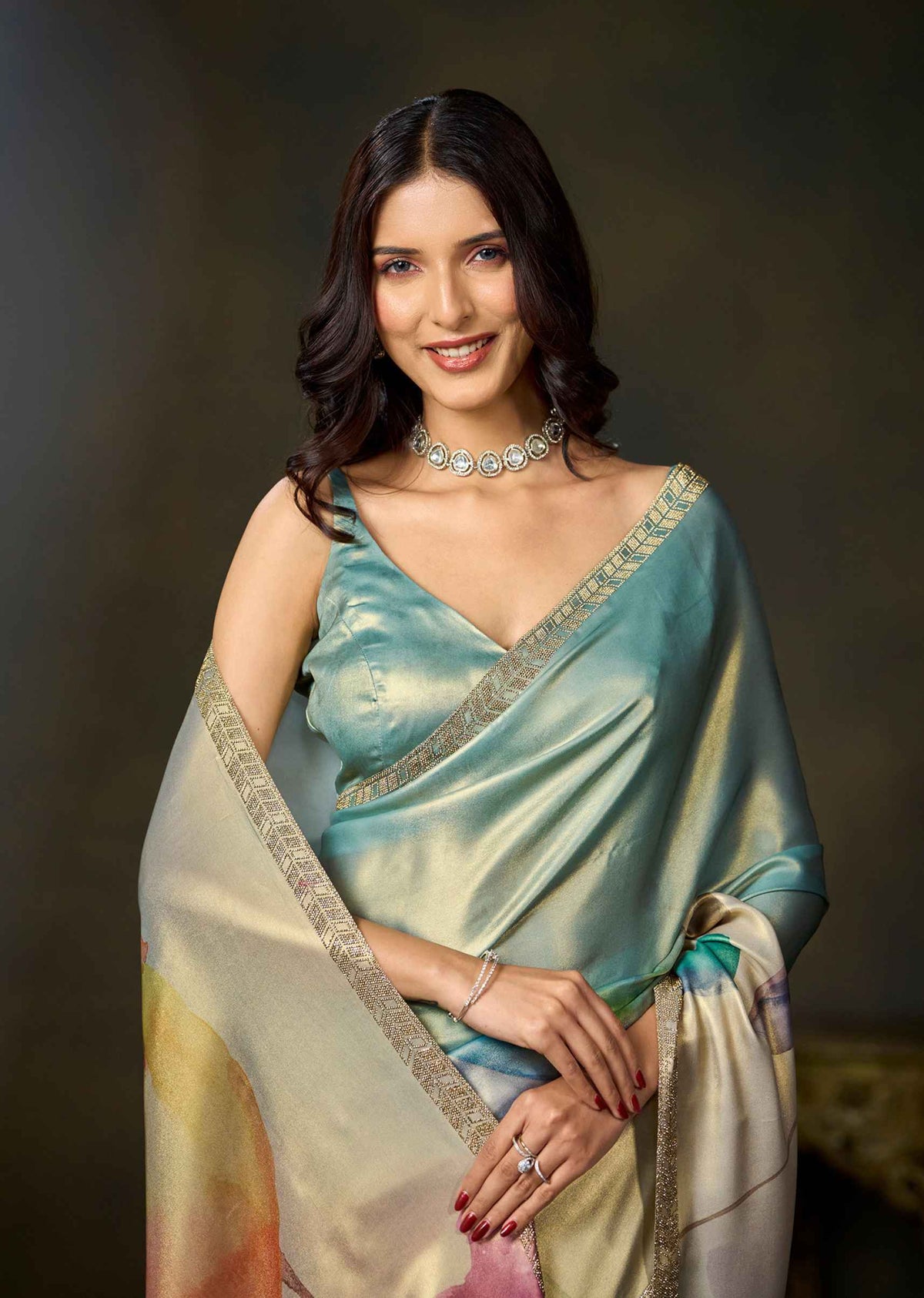A woman wearing a draped satin saree in shades of teal, cream, and gold with a metallic embroidered border, styled with a matching embellished blouse and jewelry against a dark studio background.