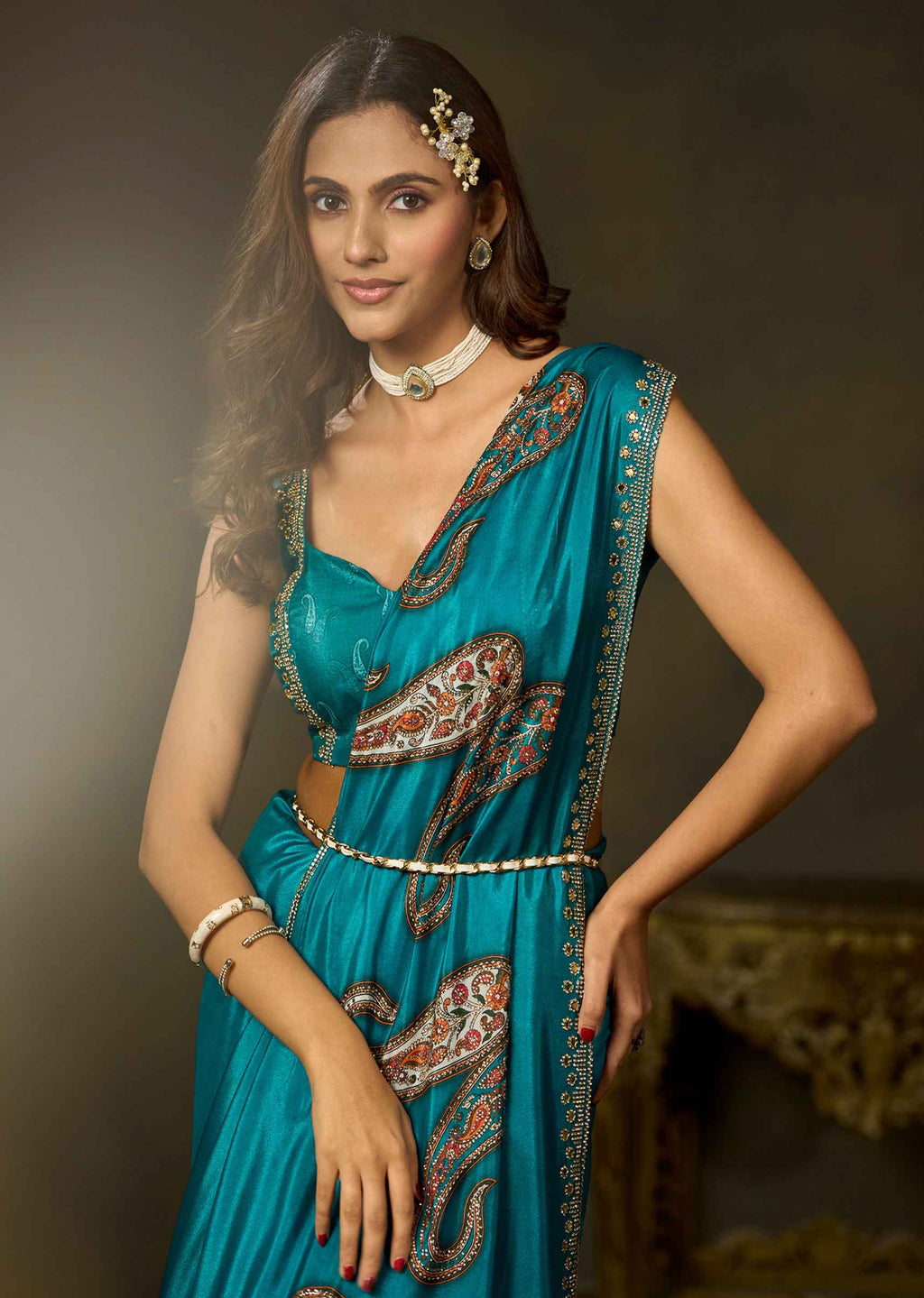 A woman modeling a teal blue sari with intricate red, gold, and white paisley patterns, styled with a matching belt and jewelry against a dark brown studio background.