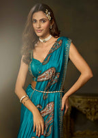 A woman modeling a teal blue sari with intricate red, gold, and white paisley patterns, styled with a matching belt and jewelry against a dark brown studio background.