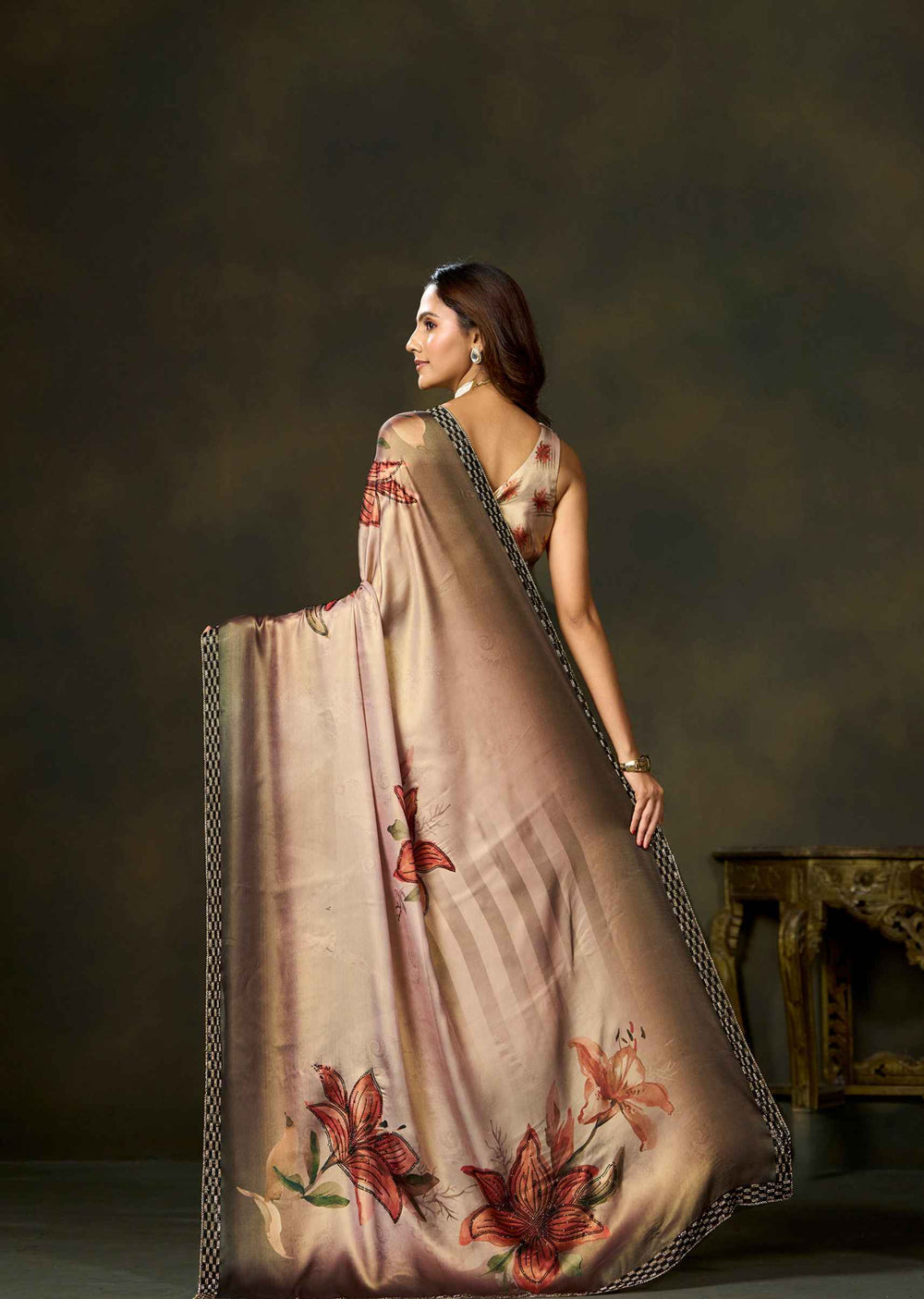 A woman wearing a draped beige saree with red floral motifs and a dark patterned border, photographed in a studio setting against a dark brown background.
