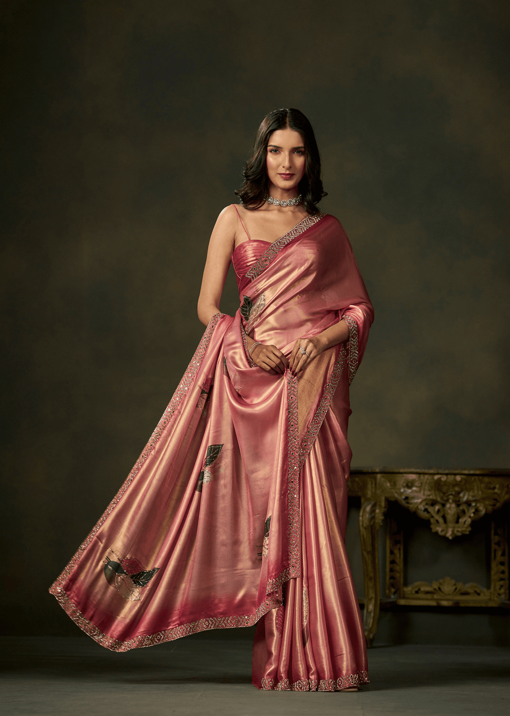 A woman models a pink satin saree with black floral embroidery and a metallic gold border, posed against a dark brown studio backdrop with an ornate table in the background.
