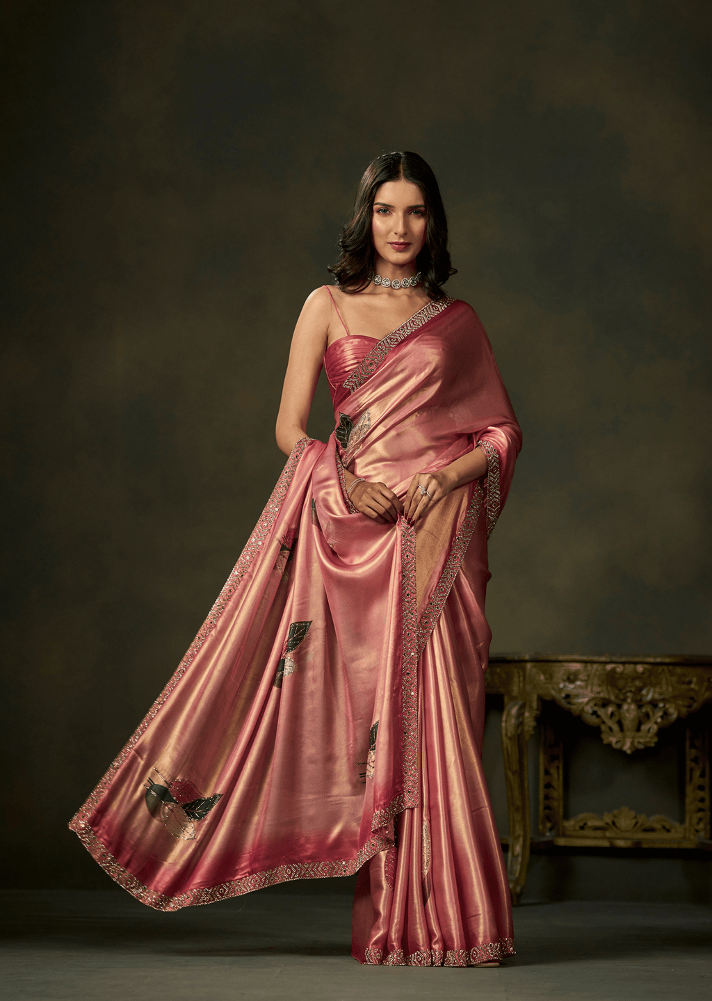 A woman models a pink satin saree with black floral embroidery and a metallic gold border, posed against a dark brown studio backdrop with an ornate table in the background.