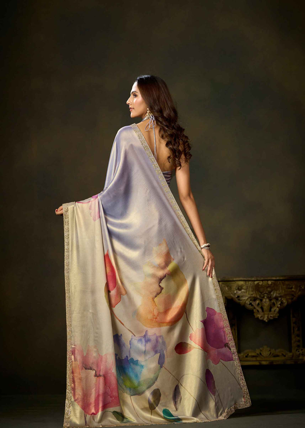 A woman wearing a draped satin saree with a pastel lavender body and a watercolor floral print in pink, blue, and orange, posed against a dark studio background.
