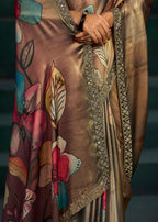 A close-up of a draped satin saree with a metallic bronze-brown base, decorated with colorful floral motifs in pink, teal, and yellow, and finished with an ornate gold embroidered border.