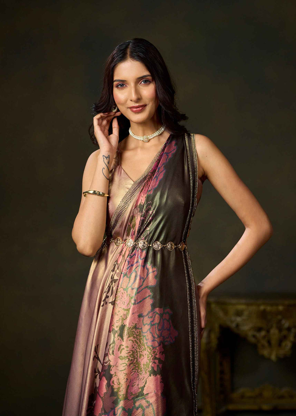 A woman models a sleeveless, V-neck dress with a metallic bronze sheen and a pink floral print, styled with gold jewelry against a dark, elegant background.