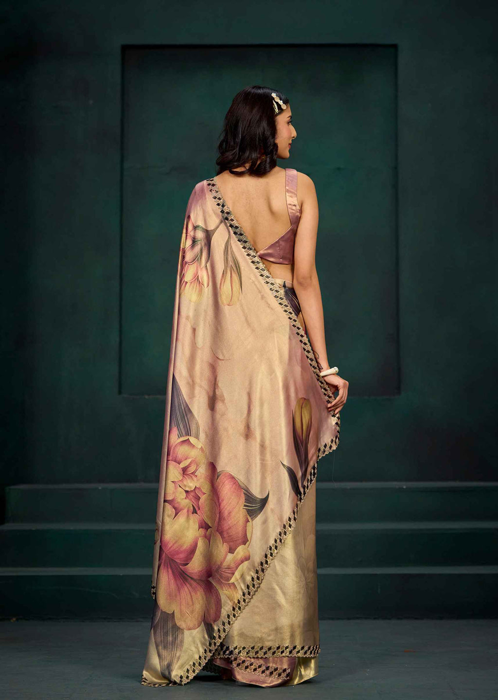 A woman stands with her back to the camera wearing a beige saree with a large floral print in pink, yellow, and green, paired with a mauve-colored blouse, set against a dark green textured wall.