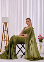 A woman seated on a black folding chair wearing an olive green saree with silver embroidered motifs, posed in front of white curtains with a wooden tripod floor lamp and a small side table with flowers.