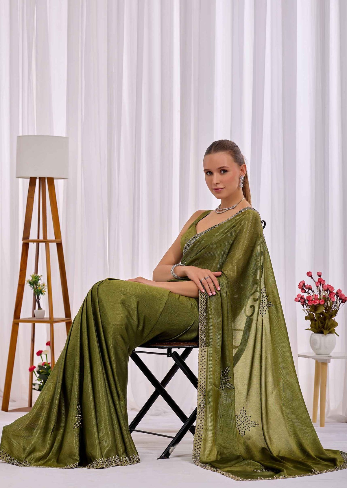 A woman seated on a black folding chair wearing an olive green saree with silver embroidered motifs, posed in front of white curtains with a wooden tripod floor lamp and a small side table with flowers.