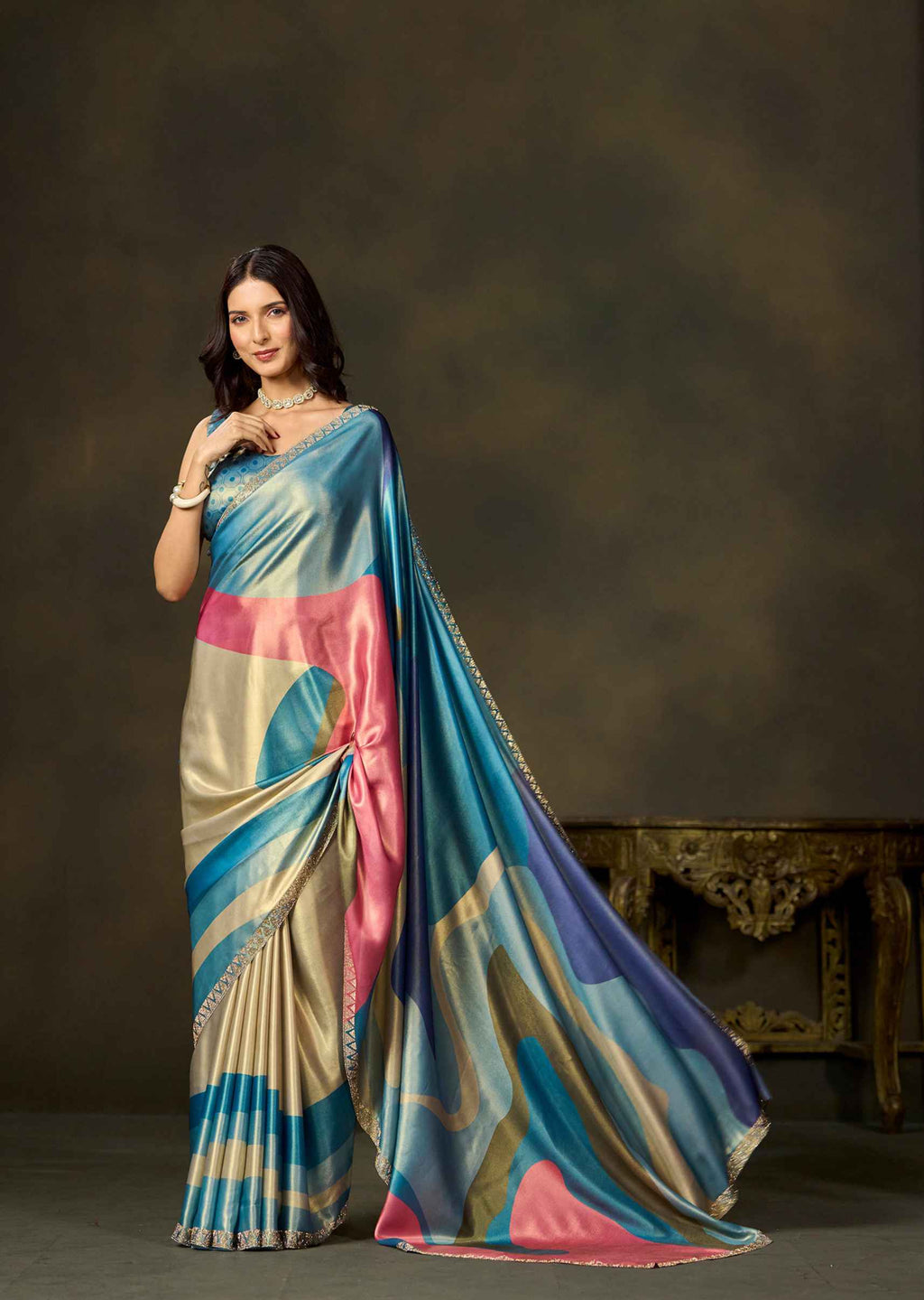 A woman models a draped saree with a multicolor abstract pattern of blue, pink, beige, and gold, set against a dark brown studio background with an ornate table.